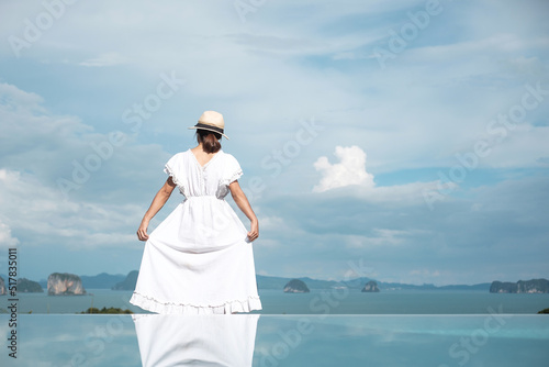 Happy traveler woman in white dress enjoy Beautiful view, alone Tourist with hat standing and relaxing over swimming pool. travel, summer and vacation concept © Jo Panuwat D