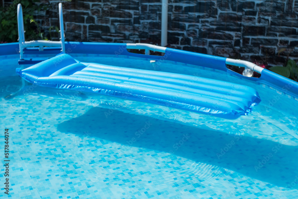 Blue inflatable mattress in a round frame pool of a country house. The pool has the purest blue water. In the background is a dark stone wall.