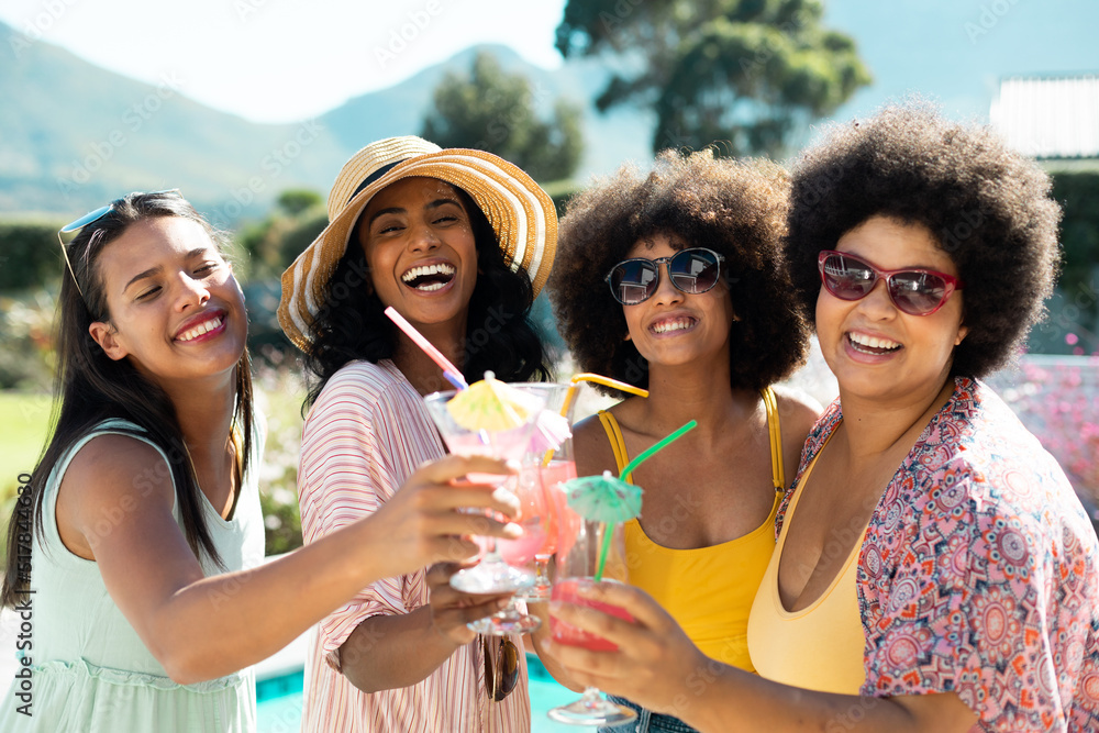 Cheerful biracial female friends toasting cocktails and posing while enjoying pool party in summer Cheerful biracial female friends toasting cocktails and posing while enjoying pool party in summer