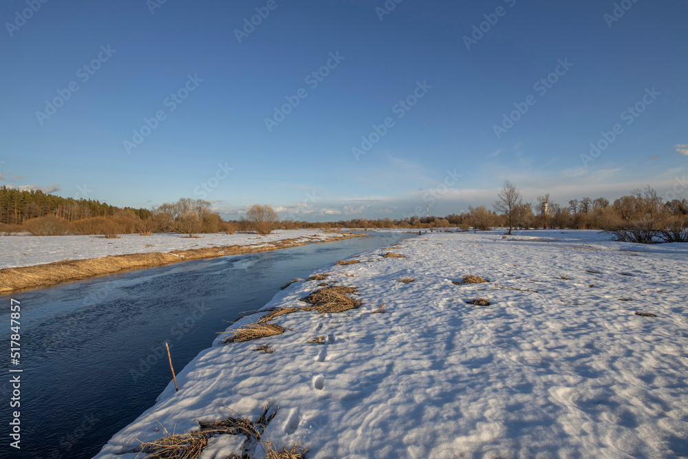A picturesque landscape, early spring, a river with snow-covered banks, dry grass and bushes. March sunny day by the river. The first thaws, the snow is melting. A picturesque landscape, early spring, a river with snow-covered banks, dry grass and bushes. March sunny day by the river. The first thaws, the snow is melting.