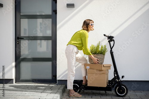 Young woman carrying basket with herbs on a porch of her house with electric scooter and parcels nearby. Concept of sustainability and modern eco-friendly lifestyle