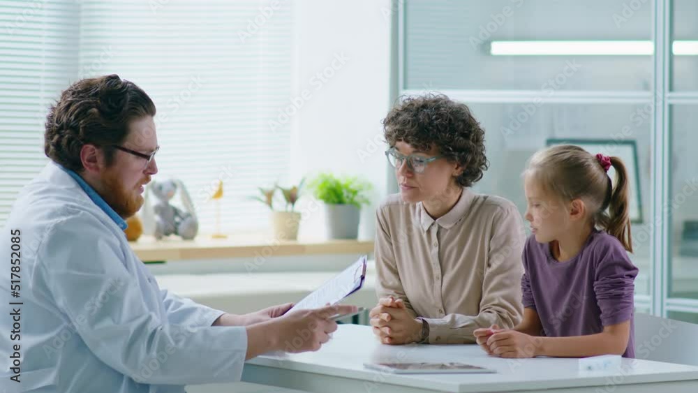 Woman sitting with little daughter in clinic, giving clipboard with medical test results to male pediatrician and speaking with him during consultation Woman sitting with little daughter in clinic, giving clipboard with medical test results to male pediatrician and speaking with him during consultation