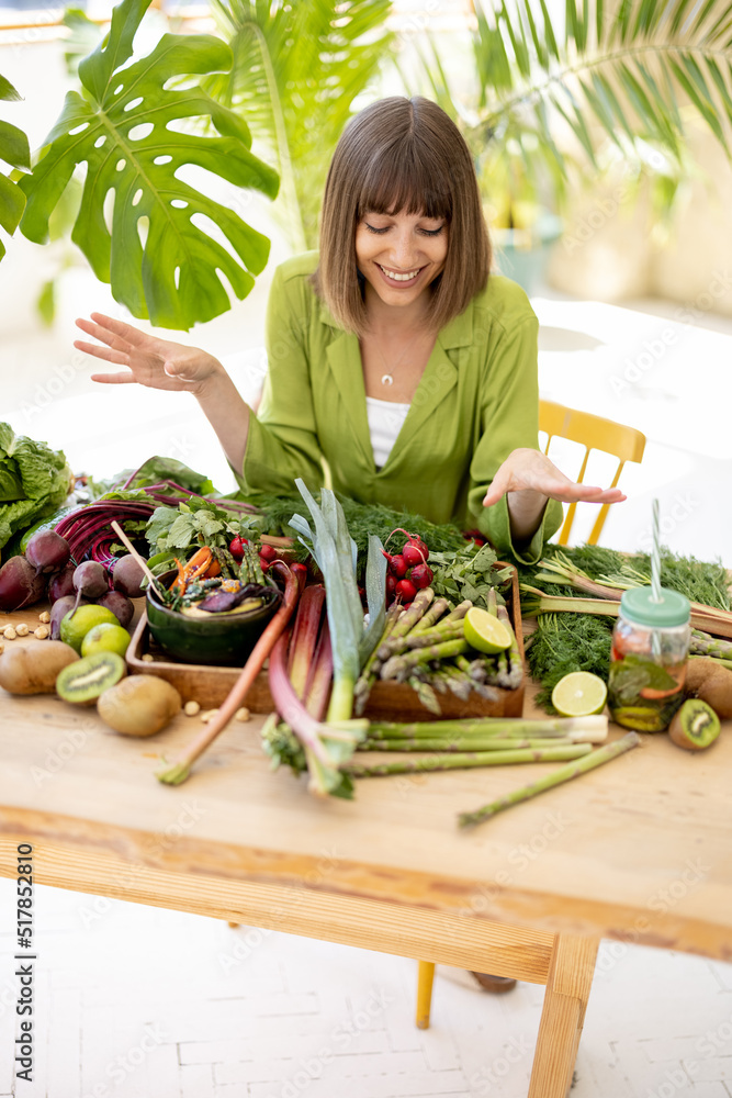 Obraz premium Portrait of a young cheerful sits by the table full of fresh vegetables, fruits and greens indoors with plants on background. Healthy eating and lifestyle concept