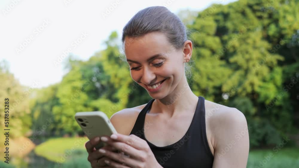 Happy beautiful woman standing in the park using mobile phone app smile sharing sportive results in social media after finishing training. Active sport girl typing on smartphone at sunset workout. Happy beautiful woman standing in the park using mobile phone app smile sharing sportive results in social media after finishing training. Active sport girl typing on smartphone at sunset workout.