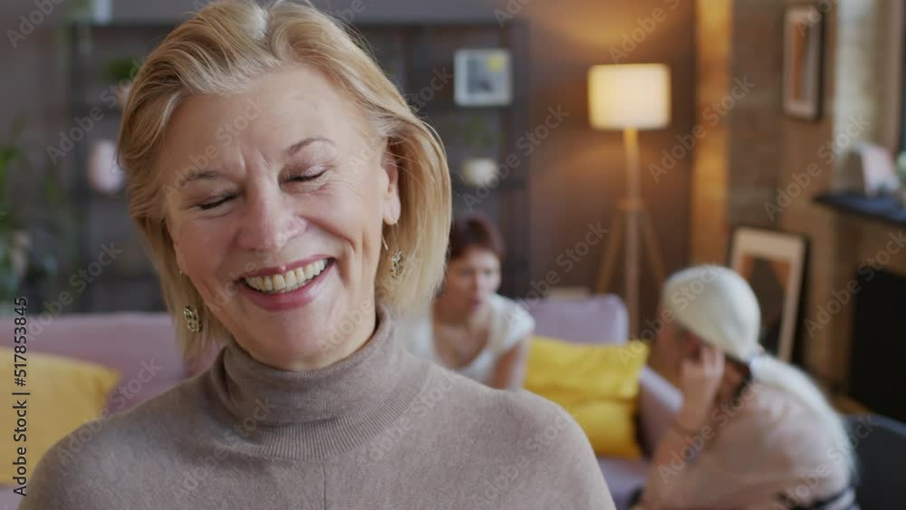 Slowmo closeup portrait of elegant mature blonde woman smiling at camera standing in modern loft style living room. Her two girlfriends chatting on sofa in background Slowmo closeup portrait of elegant mature blonde woman smiling at camera standing in modern loft style living room. Her two girlfriends chatting on sofa in background