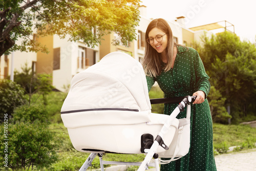 a woman in a green dress walks with a stroller. A young mother walks with a white color stroller on a sunny day. Mom looks into the stroller to the baby. photo