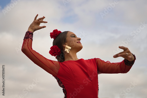 Portrait of young teenage woman in red dance suit with red carnations in her hair doing flamenco poses with clouds in the sky in the background. Flamenco concept, dance, art, typical Spanish dance. photo