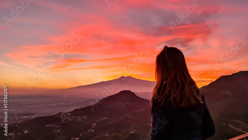 Tourist woman watching beautiful sunset behind volcano Mount Etna near Castelmola, Taormina, Sicily, Italy, Europe, EU. Clouds with vibrant red orange colors. Silhouette of person during twilight