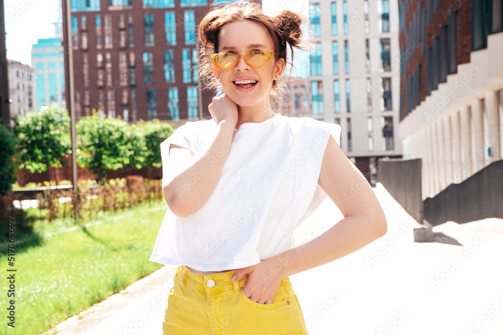 Young beautiful smiling hipster woman in trendy summer yellow jeans clothes. Carefree teen model posing in the street at sunset. Positive female outdoors. Cheerful and happy. In sunglasses