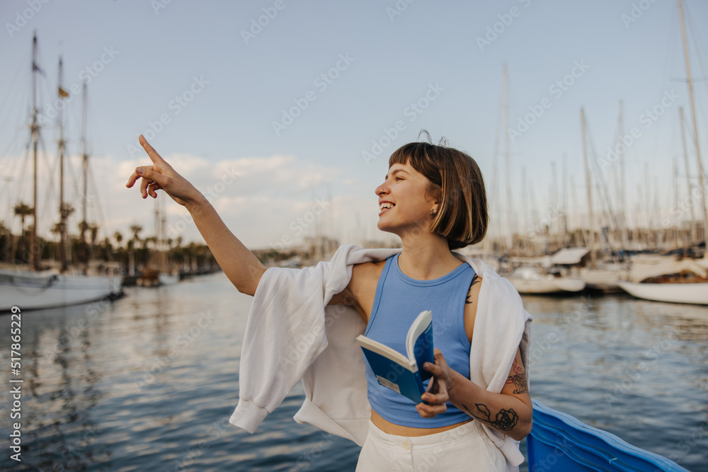 Funny young caucasian woman points finger to side spending time outdoors by sea. Brown-haired with bob haircut wears blue T-shirt, white sweatshirt on top. Sincere emotions lifestyle concept