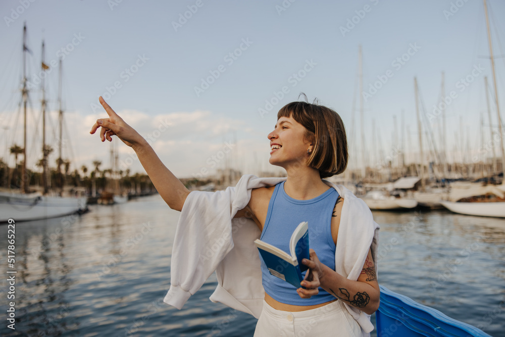 Funny young caucasian woman points finger to side spending time outdoors by sea. Brown-haired with bob haircut wears blue T-shirt, white sweatshirt on top. Sincere emotions lifestyle concept Funny young caucasian woman points finger to side spending time outdoors by sea. Brown-haired with bob haircut wears blue T-shirt, white sweatshirt on top. Sincere emotions lifestyle concept