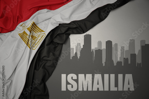 abstract silhouette of the city with text Ismailia near waving colorful national flag of egypt on a gray background. photo