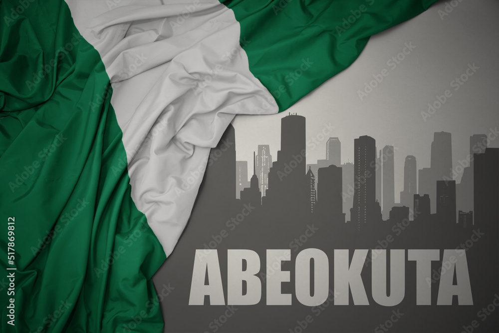 abstract silhouette of the city with text Abeokuta near waving colorful national flag of nigeria on a gray background.