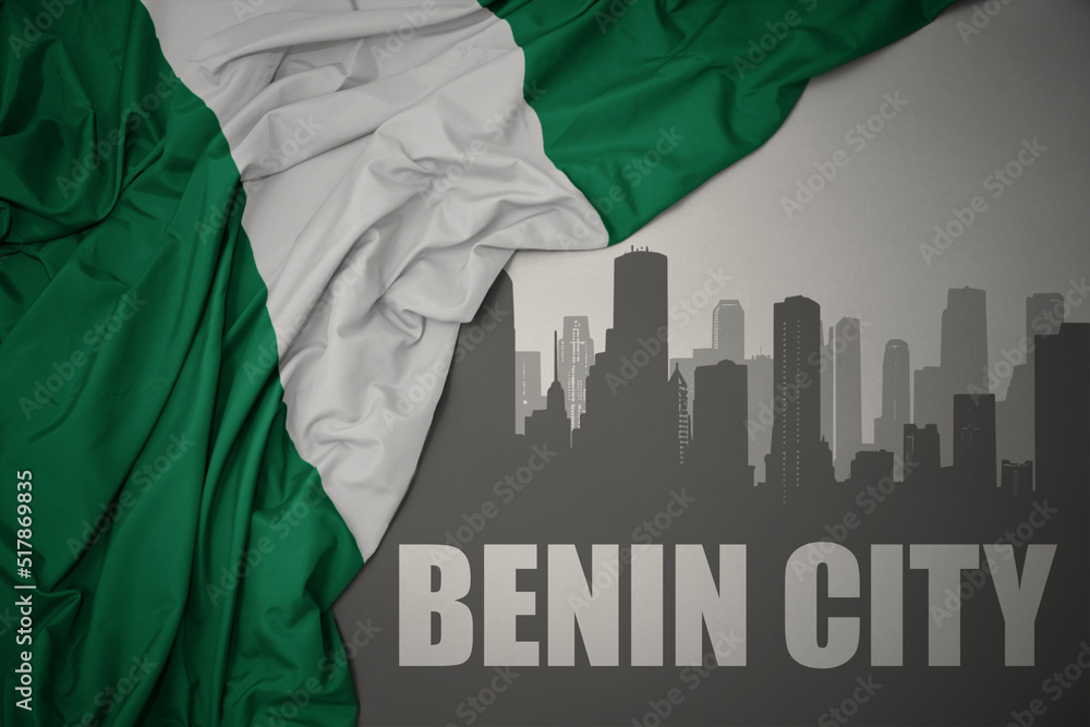 abstract silhouette of the city with text Benin City near waving colorful national flag of nigeria on a gray background.