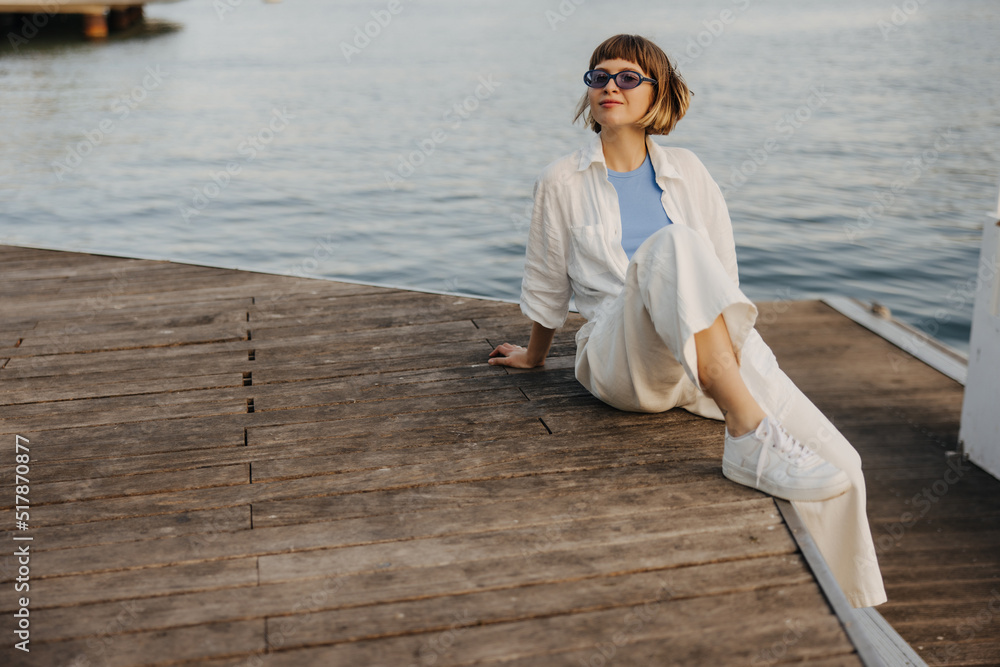 Stylish young caucasian woman in good weather is resting sitting on pier by sea. Brown-haired with bob haircut wears casual clothes. Summer vacation concept