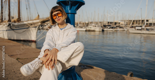 Beautiful caucasian young lady in white tracksuit sits on pier near sea during day. Brown-haired woman with bob haircut wears sunglasses. Vacation, happiness and pleasure concept