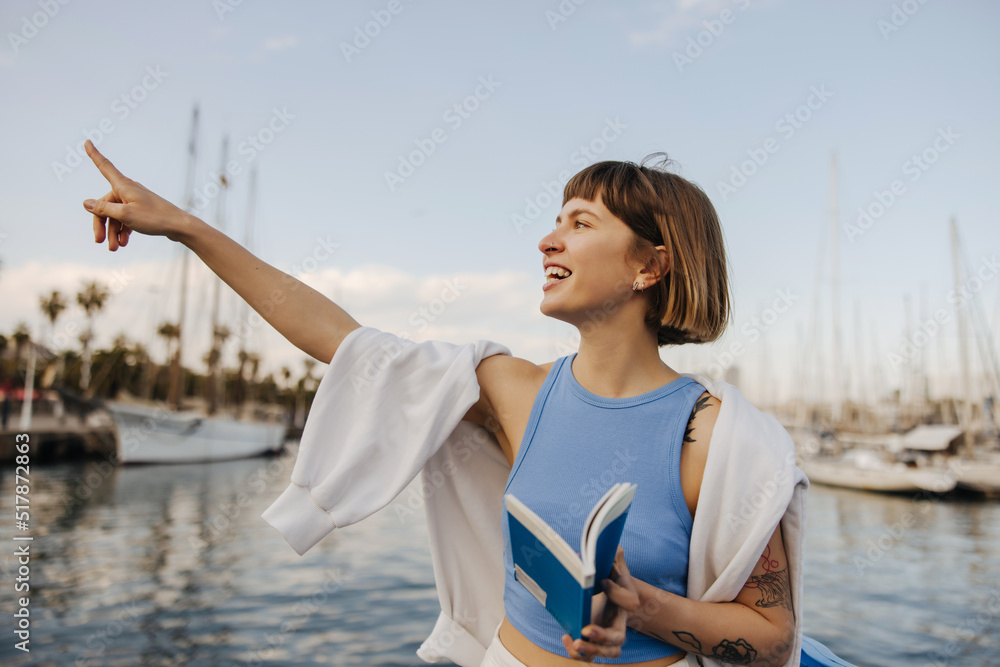 Cheerful young caucasian lady points her finger to sky, standing near sea on warm summer day. Brown-haired woman with bob haircut wears T-shirt and sweatshirt on top. Sincere emotions concept Cheerful young caucasian lady points her finger to sky, standing near sea on warm summer day. Brown-haired woman with bob haircut wears T-shirt and sweatshirt on top. Sincere emotions concept