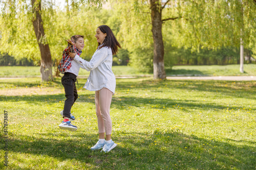 Cute little boy jumping and having fun with pretty cheerful mother in green park. Outdoors.