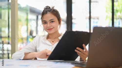 Pleasant young businesswoman holding clipboard, working online on laptop at her workplace