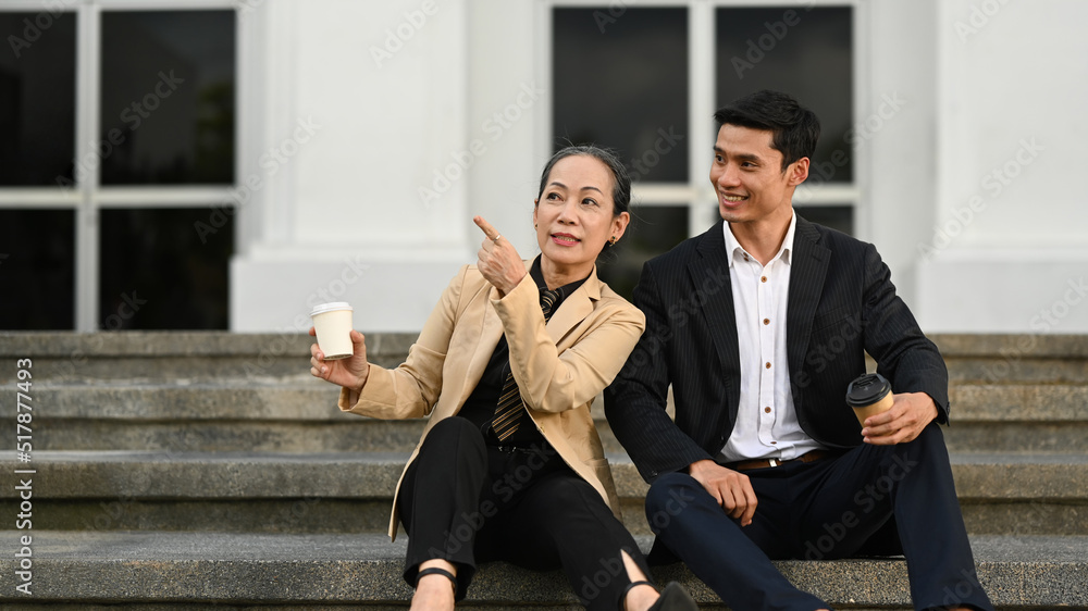 Mature businesswoman and young businessman talking each other, sitting on the stairs in front of business center Mature businesswoman and young businessman talking each other, sitting on the stairs in front of business center