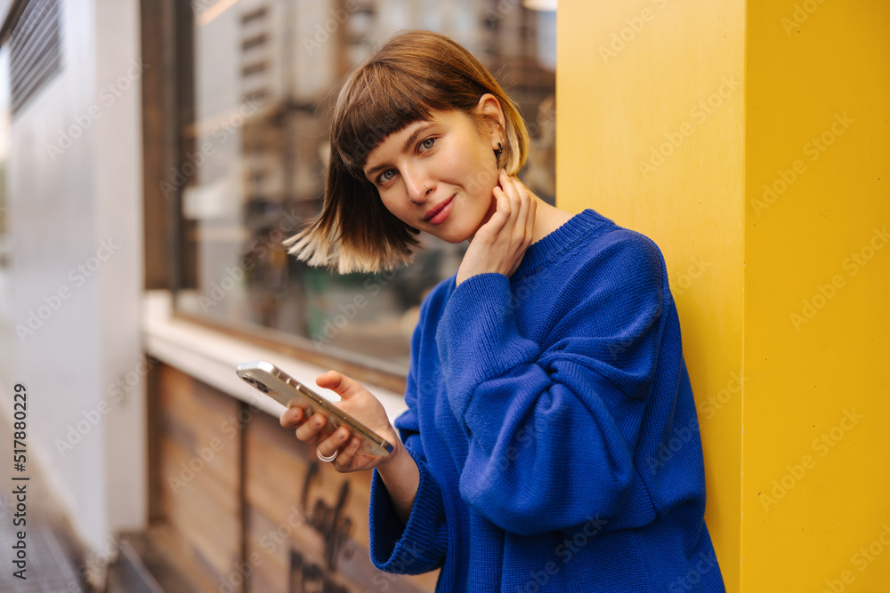 Pretty young caucasian brown-haired woman looking at camera using smartphone outdoors. Short-haired lady wears blue sweatshirt. Concept of city life, technology Pretty young caucasian brown-haired woman looking at camera using smartphone outdoors. Short-haired lady wears blue sweatshirt. Concept of city life, technology