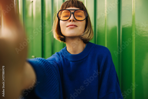Foto Cute young caucasian girl makes selfie looking at camera on background of green wall street