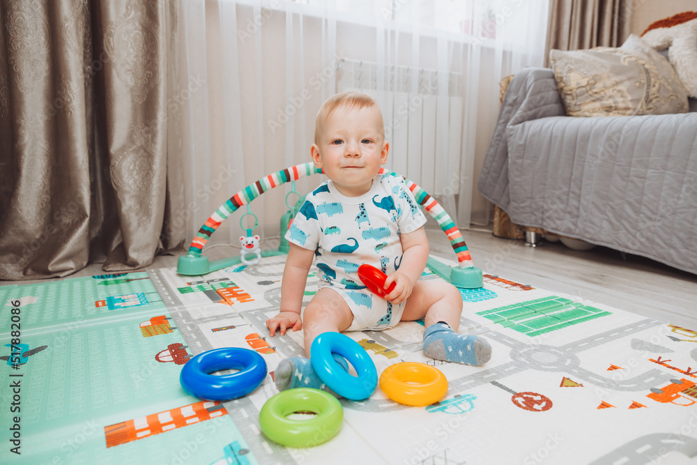 A cute baby is playing with a rainbow toy pyramid while sitting on a play mat in a sunny bedroom. Toys for small children. A cute baby is playing with a rainbow toy pyramid while sitting on a play mat in a sunny bedroom. Toys for small children.