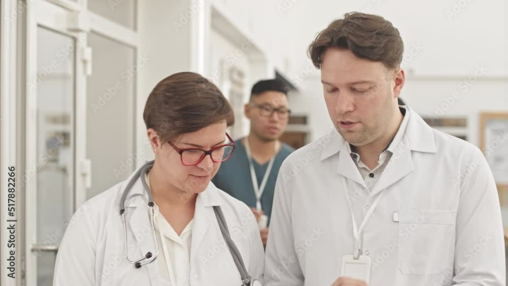 Medium slowmo of couple of doctors talking while walking along modern hospital corridor. Two multiethnic male nurses in scrubs walking behind them having conversation with each other Medium slowmo of couple of doctors talking while walking along modern hospital corridor. Two multiethnic male nurses in scrubs walking behind them having conversation with each other