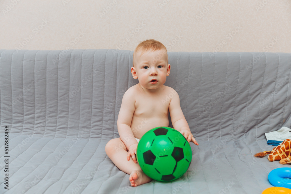 the kid is holding a soccer ball, sitting on the couch barefoot and looking away. Adorable baby is playing at home on the couch. The concept of holidays, weekends and childhood the kid is holding a soccer ball, sitting on the couch barefoot and looking away. Adorable baby is playing at home on the couch. The concept of holidays, weekends and childhood