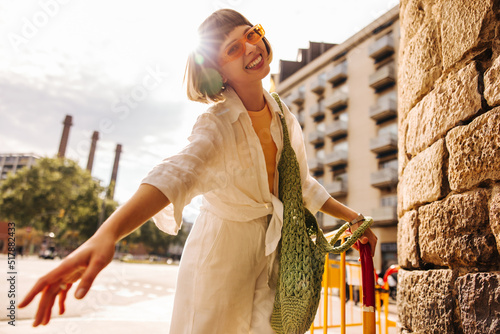 Cheerful young caucasian girl smiles with teeth at camera enjoying summer weekend. Fair-haired lady wears sunglasses, shirt, pants and bag. City life concept