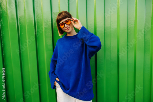 Fashionable young caucasian woman in sunglasses posing against backdrop of green city wall. Brown-haired with bob haircut wears blue sweatshirt. Lifestyle concept