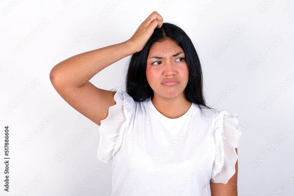 young brunette woman wearing white T-shirt standing against white background saying: Oops, what did I do? Holding hand on head with frightened and regret expression.
