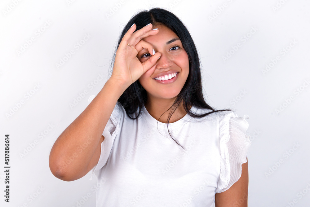 young brunette woman wearing white T-shirt standing against white background with happy face smiling doing ok sign with hand on eye looking through finger. young brunette woman wearing white T-shirt standing against white background with happy face smiling doing ok sign with hand on eye looking through finger.