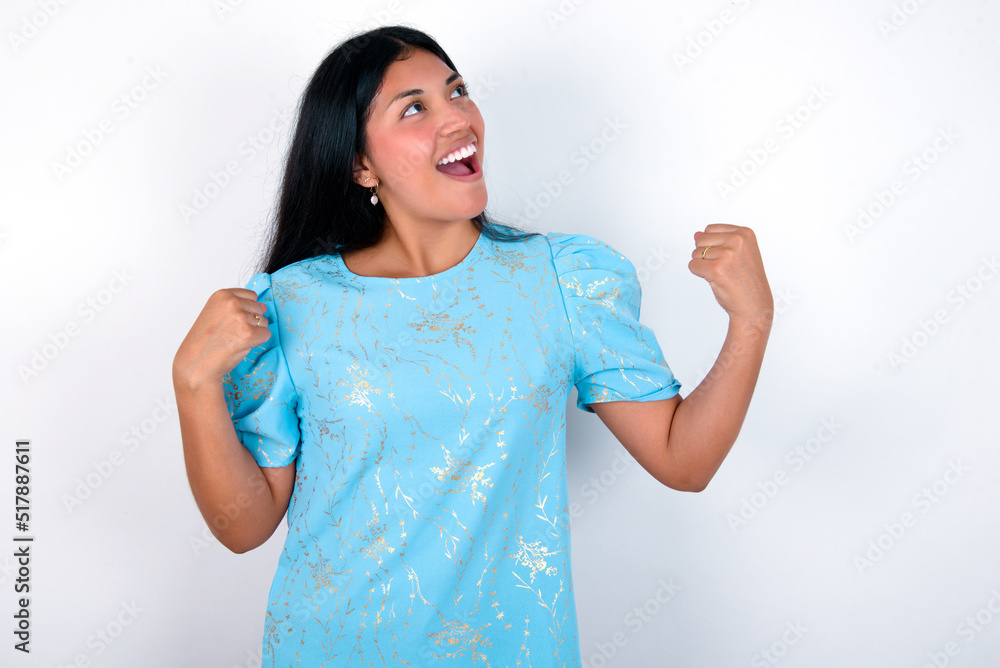 Attractive young brunette woman wearing blue T-shirt standing against white background celebrating a victory punching the air with his fists and a beaming toothy smile Attractive young brunette woman wearing blue T-shirt standing against white background celebrating a victory punching the air with his fists and a beaming toothy smile