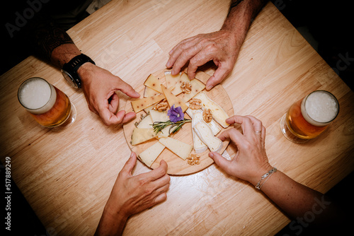 four hands picking up pieces of cheese presented on a wooden board on a table with two beers