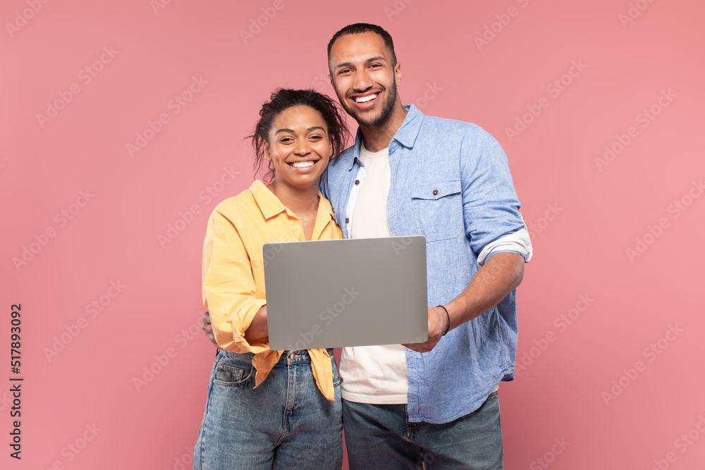 Portrait of happy african american couple holding and using laptop, posing on pink background and smiling at camera Portrait of happy african american couple holding and using laptop, posing on pink background and smiling at camera