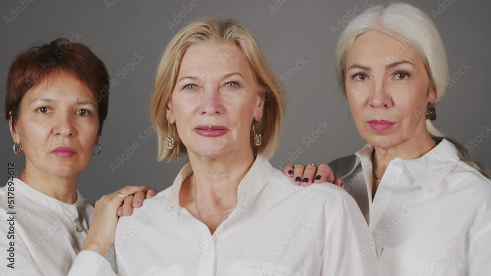 Chest up studio portrait of three confident mature women in white shirts posing for camera standing on gray background Chest up studio portrait of three confident mature women in white shirts posing for camera standing on gray background