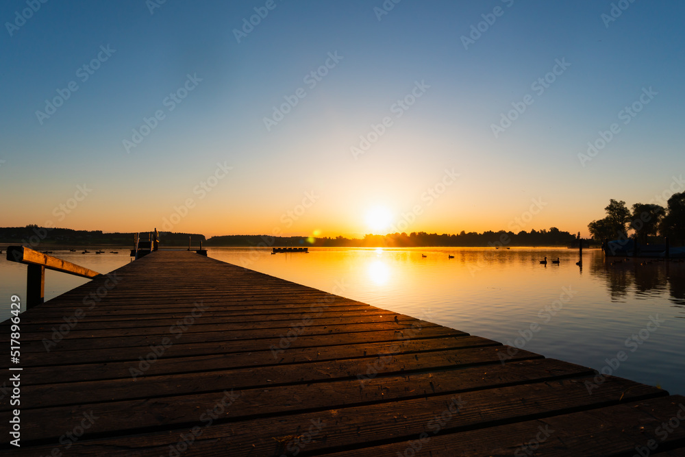 chill sunrise with a jetty close up in foreground, forest and a few ducks as silhouettes, complete blue sky and no clouds
