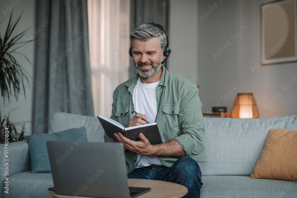 Smiling happy aged european man with beard in headphones has meeting remotely at computer Smiling happy aged european man with beard in headphones has meeting remotely at computer
