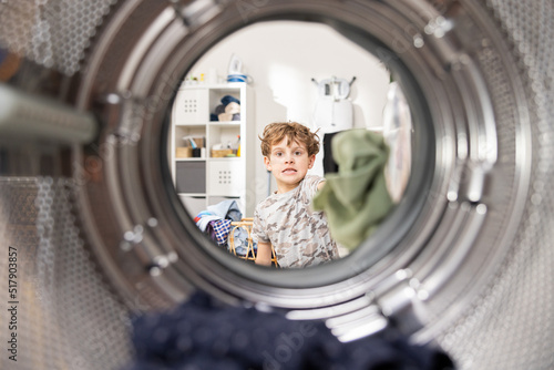 Small cheerful boy interested in the operation of the device puts clothes into the drum of the washing machine, helps his mother in household duties, resourceful, hard-working child.