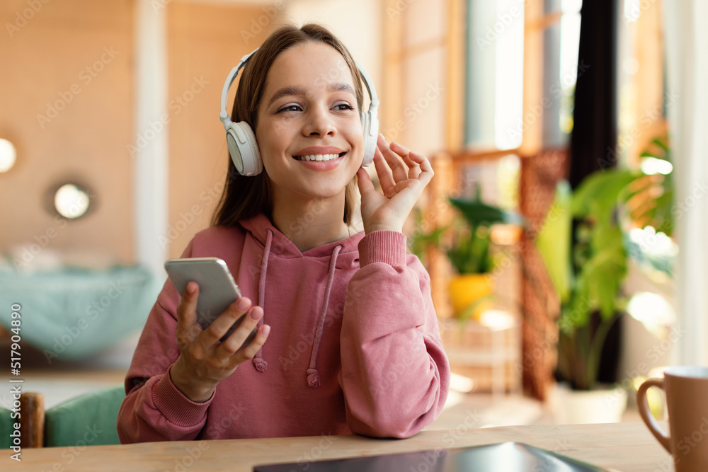 Happy caucasian teen girl wearing wireless headphones and using cellphone, sitting at desk at home and smiling Happy caucasian teen girl wearing wireless headphones and using cellphone, sitting at desk at home and smiling