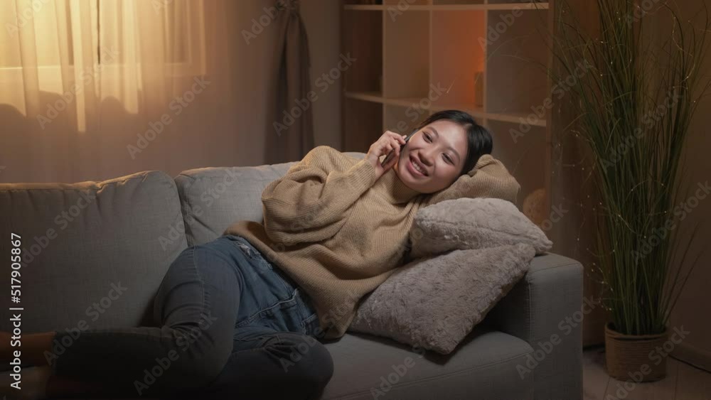 Home phone call. Evening rest. Mobile communication. Relaxed smiling girl enjoying pleasant conversation lying on cozy couch in modern living room interior with free space. Home phone call. Evening rest. Mobile communication. Relaxed smiling girl enjoying pleasant conversation lying on cozy couch in modern living room interior with free space.