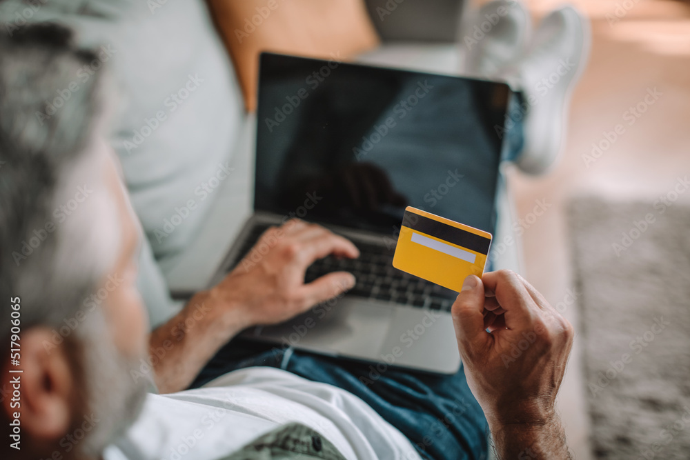 Mature european male resting on sofa and shopping online with credit card and laptop with blank screen Mature european male resting on sofa and shopping online with credit card and laptop with blank screen