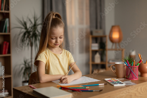 Little Girl Drawing With Paintbrush Sitting At Desk At Home