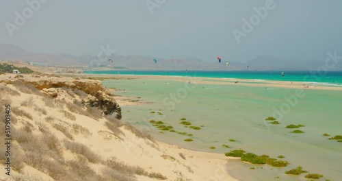 Group of unrecognizable kitesurfers kiteboarders tourists enjoy hobby. Wide shot of nature white beach landscape and blue shallow ocean water. Watersport on Canary Islands, Fuerteventura, Spain. photo