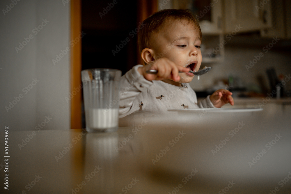A one-year-old child sits at home at the kitchen table and has breakfast, he eats porridge on his own and drinks milk from a glass. A one-year-old child sits at home at the kitchen table and has breakfast, he eats porridge on his own and drinks milk from a glass.