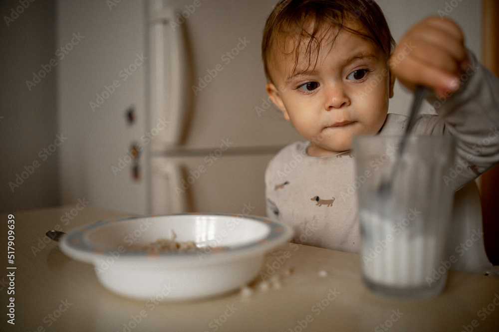 A one-year-old child sits at home at the kitchen table and has breakfast, he eats porridge on his own and drinks milk from a glass. A one-year-old child sits at home at the kitchen table and has breakfast, he eats porridge on his own and drinks milk from a glass.