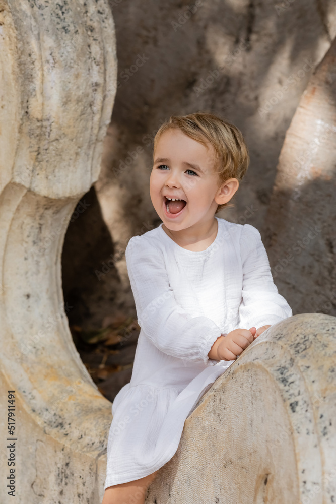 Excited toddler kid in white dress sitting on stone bench in park in Spain. Excited toddler kid in white dress sitting on stone bench in park in Spain.