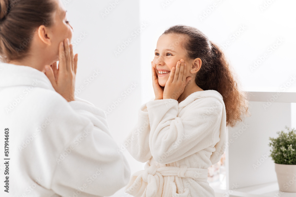beauty, hygiene, morning and people concept - happy smiling mother and daughter touching their faces in bathroom