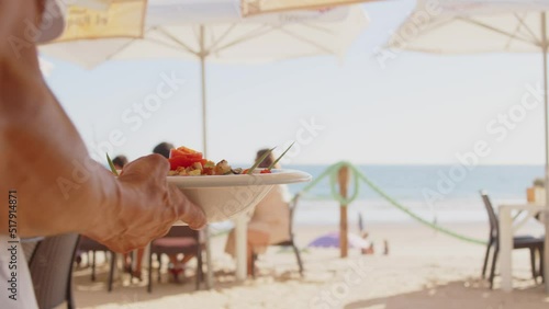 4K SlowMo - professional waiter bringing a plate of salad to a family's table in a beachside restaurant